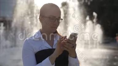 白人年轻女子戴着光学眼镜出现在公园喷泉附近，手里拿着智能手机。